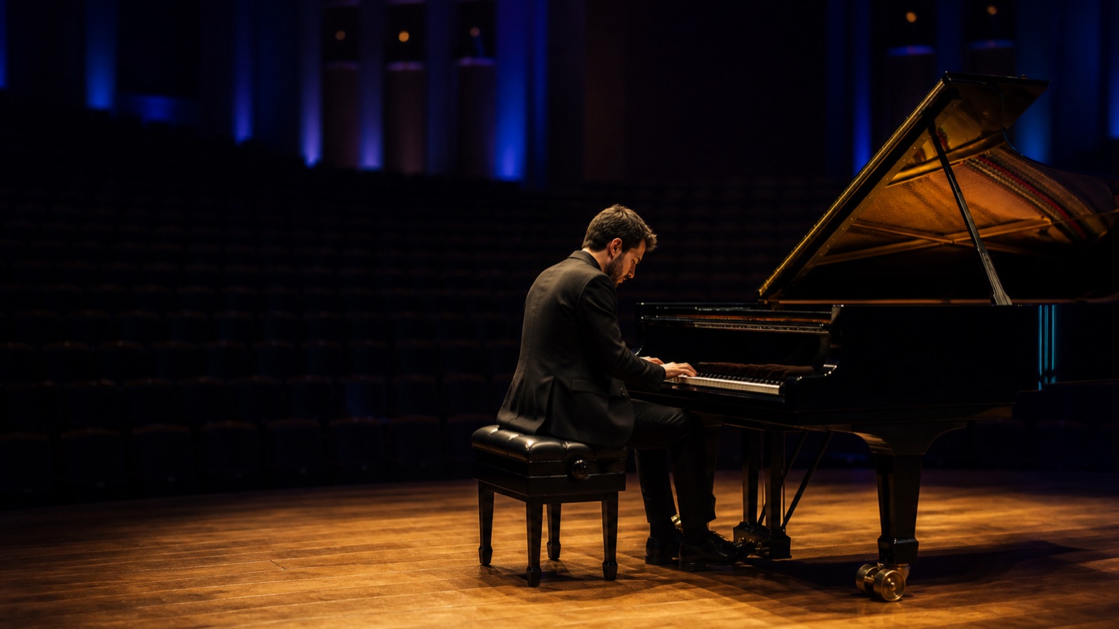 A pianist rehearses at a grand piano in a warmly lit concert hall.
