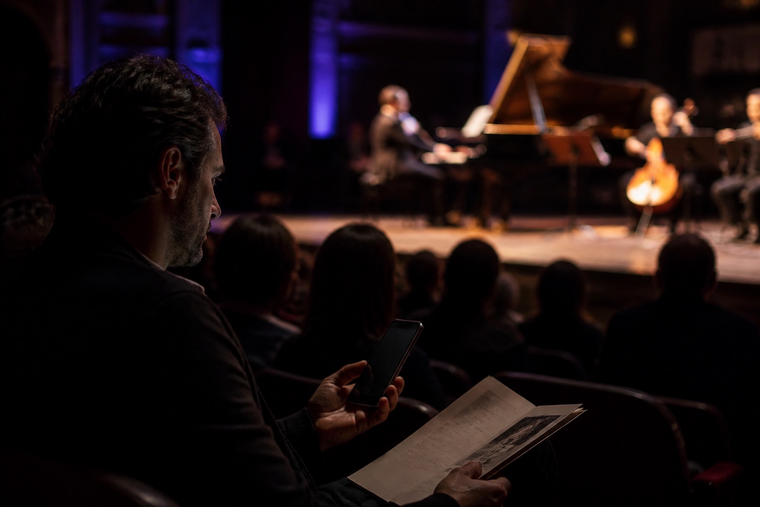 A listener sits near a live performance while holding an unbranded phone with a dark screen.